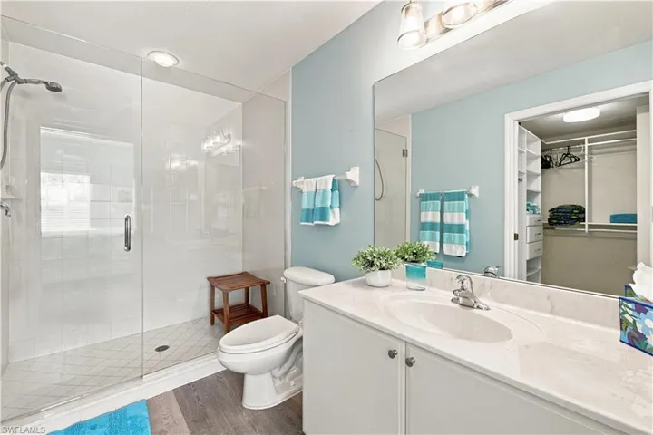 Glass-enclosed shower with white tiled walls, wood-finish flooring, white vanity with integrated sink, and large wall-mounted mirror