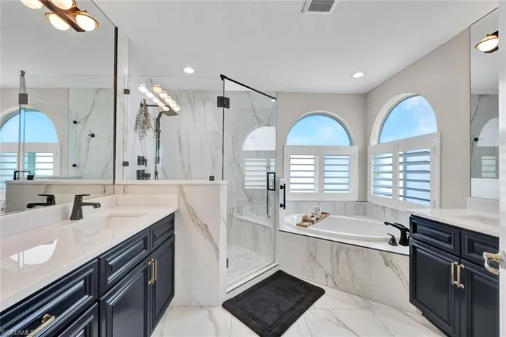 Dual vanities with undermount sinks, a built-in soaking tub, an enclosed glass shower, and arched windows with fogged glass and plantation shutters