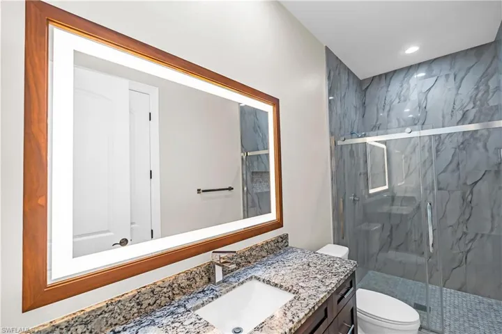 Bathroom featuring an illuminated wood-framed mirror, a stone-finish countertop with an undermount sink, a dark wood-finish vanity, a glass-enclosed shower with textured wall tile, and a patterned shower floor