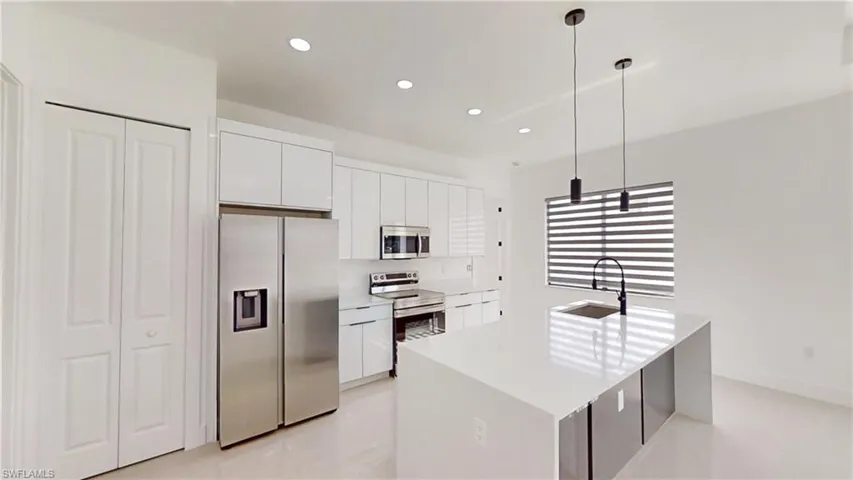 Kitchen with stainless steel appliances, a center island with sink, white cabinets, hanging light fixtures, and light stone counters