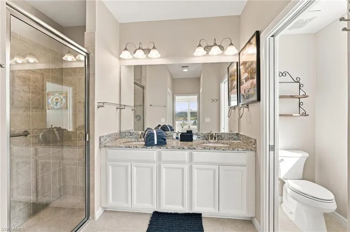 Dual-sink vanity with stone-finish countertop, white shaker-style cabinetry, full-width mirror, tile-surround shower with glass enclosure, and two three-light vanity fixtures