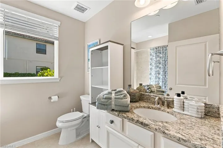 Bathroom featuring stone-surface vanity countertop, undermount sink, built-in white cabinetry with open shelving, large wall-mounted mirror, and horizontal window blinds