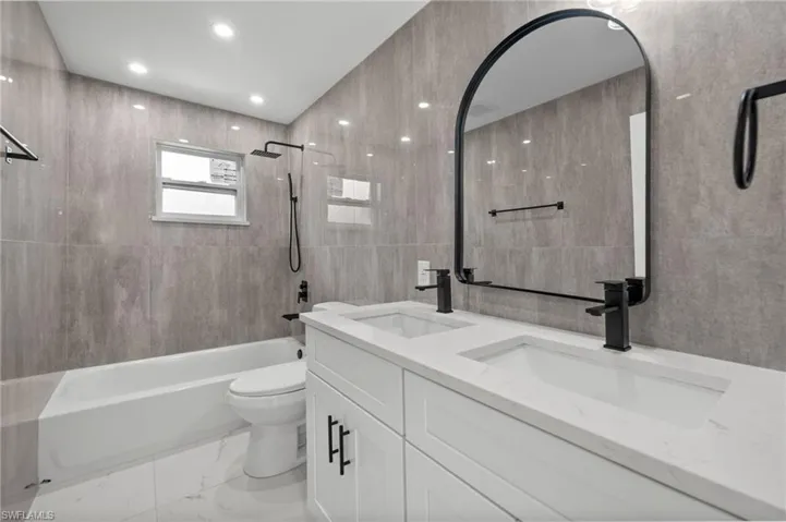 Full bath featuring bathtub / shower combination, double vanity, tile walls, light marble finish floors, and recessed lighting