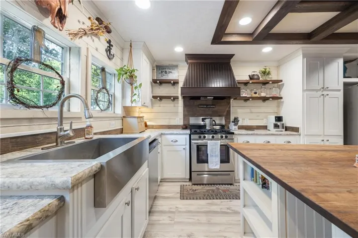 Stainless steel apron front sink with a pull-down faucet, white perimeter cabinetry, light-colored countertops, vinyl flooring, and a coffered ceiling with recessed lighting