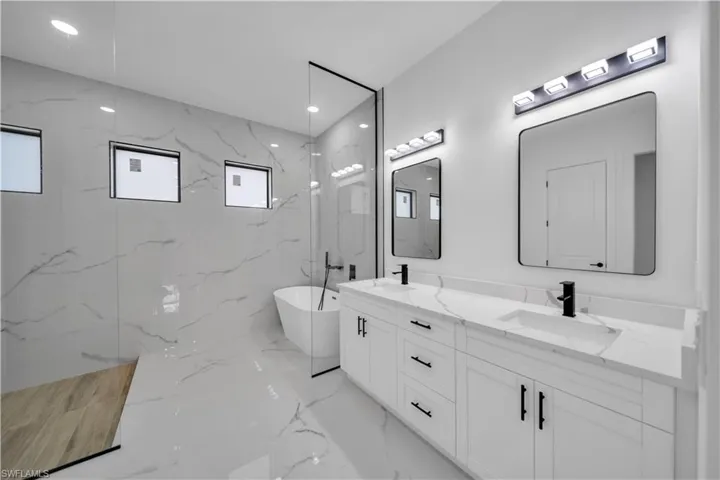 Spacious bathroom featuring a double vanity with white cabinetry, a freestanding bathtub, a glass-enclosed shower, and large format wall and floor tiling