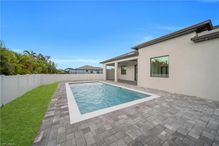 Outdoor living space featuring a rectangular swimming pool with a white coping, surrounded by grey paver decking, a manicured lawn, and a white privacy fence
