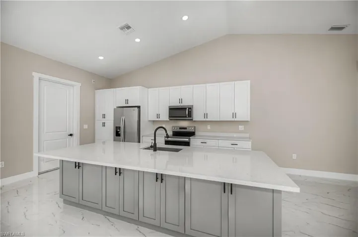 Dual tone kitchen featuring stainless steel appliances, light stone counters, a large island with sink, vaulted ceiling, and recessed lighting