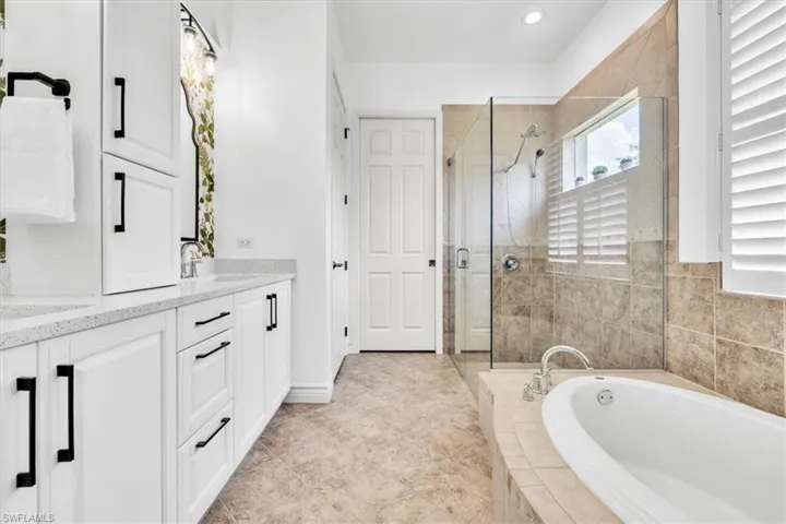 Spacious bathroom featuring a dual vanity with white cabinetry and light-toned countertops, a glass-enclosed shower with tile surround, a separate soaking tub, and louvered window treatments