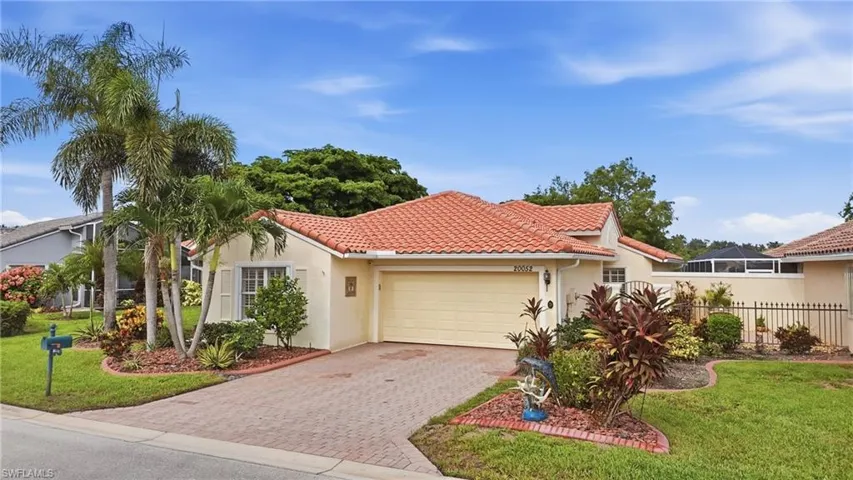 Mediterranean-style exterior with a terracotta tile roof, attached garage, paver driveway, mature palm trees, and established landscaping