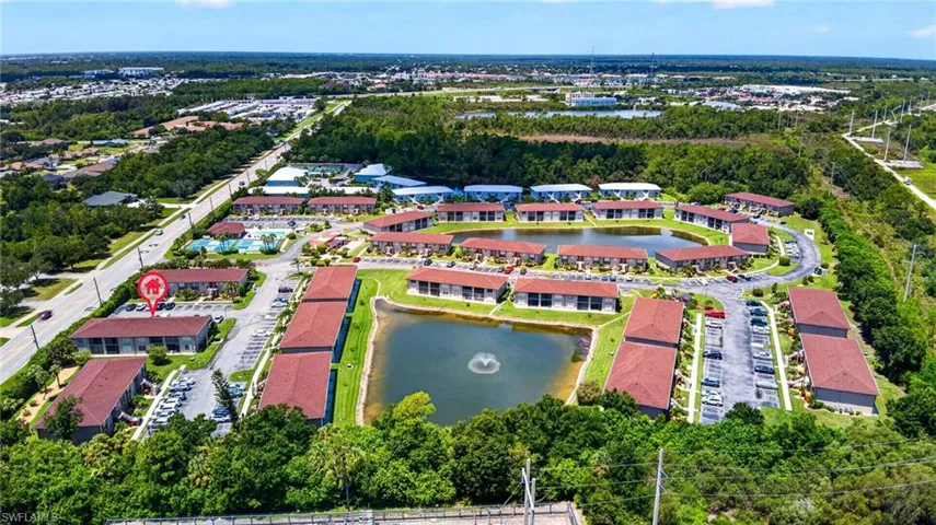 Community setting with multiple buildings featuring tiled roofs, surrounding a central pond with a fountain, and ample surface parking