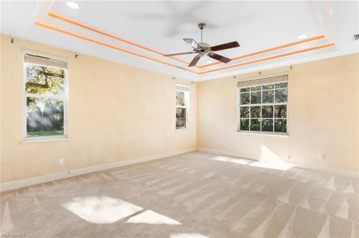 Room featuring a tray ceiling with recessed lighting and accent trim, a ceiling fan, light-toned carpet flooring, and multiple windows with blinds