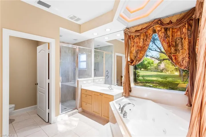 Spacious bathroom featuring a soaking tub, separate glass-enclosed shower, dual vanity with light wood cabinetry, recessed lighting, and a large window overlooking a landscaped expanse