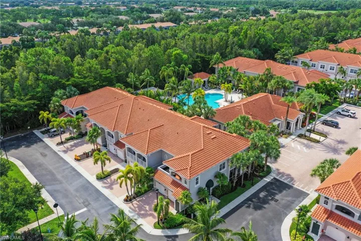 Residential complex featuring multiple buildings with terracotta-style tile roofs, a central swimming pool with surrounding lounge areas, and paved parking facilities