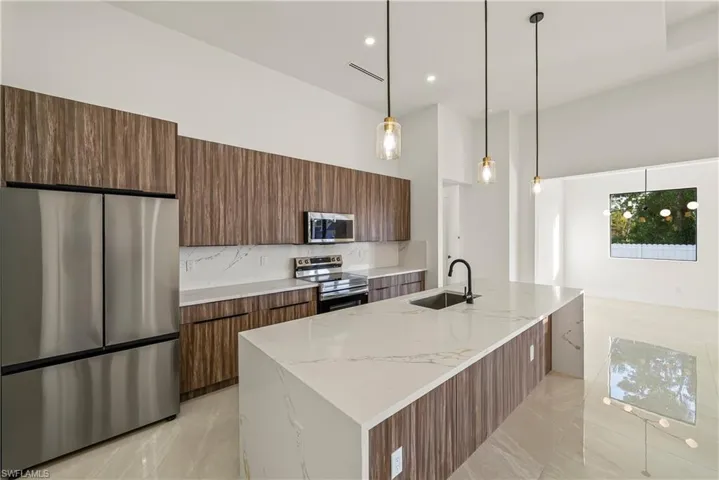 Kitchen featuring wood finish cabinetry, stainless steel appliances, a kitchen island with sink, light stone counters, and hanging light fixtures