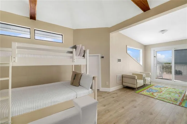 Spacious room featuring wood-finish flooring, a vaulted ceiling with exposed wood beams, and a sliding glass door opening to an outdoor balcony with a metal railing - Virtually Edited Image