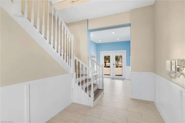 Entryway featuring a white banister staircase with wood-finish treads, a white wainscoting wall, and light-toned tile flooring - Virtually Edited Image