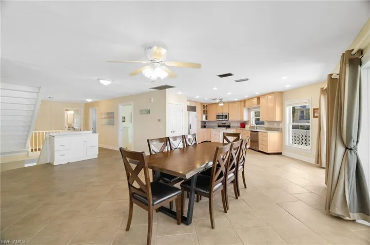 Open-concept interior featuring light-toned tile flooring, a staircase with white railing, and a kitchen with wood-finish cabinetry, stainless steel appliances, and a recessed lighting package - Virtually Edited Image