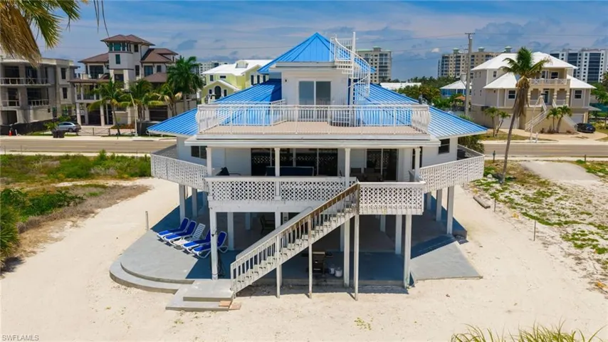 Elevated coastal residence featuring a vibrant blue metal roof, multiple levels of outdoor decking, a spiral staircase, and decorative white railings - Virtually Edited Image