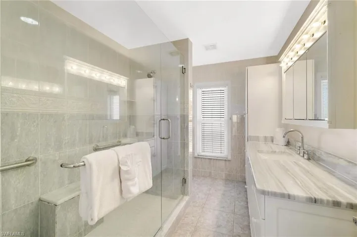Spacious bathroom featuring a glass-enclosed shower with integrated bench seating, a dual vanity with a patterned countertop, contemporary light fixtures, and tiled walls and flooring - Virtually Edited Image