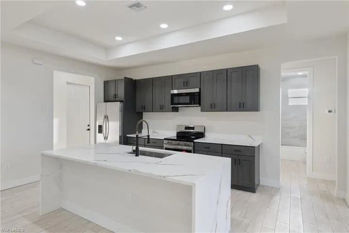 Kitchen with stainless steel appliances, light stone counters, a raised ceiling, a center island with sink, and light wood finished floors