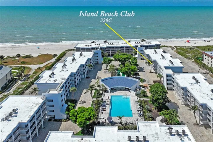 Island Beach Club 320C features a beachfront location with direct oceanfront access, a resort-style heated swimming pool surrounded by mature landscaping, contemporary multi-story residential architecture with clean lines and white exterior finishes, and expansive grounds with hardscaped amenities and green space