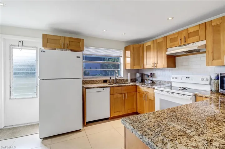 Kitchen with white appliances, under cabinet range hood, light stone countertops, backsplash, and recessed lighting