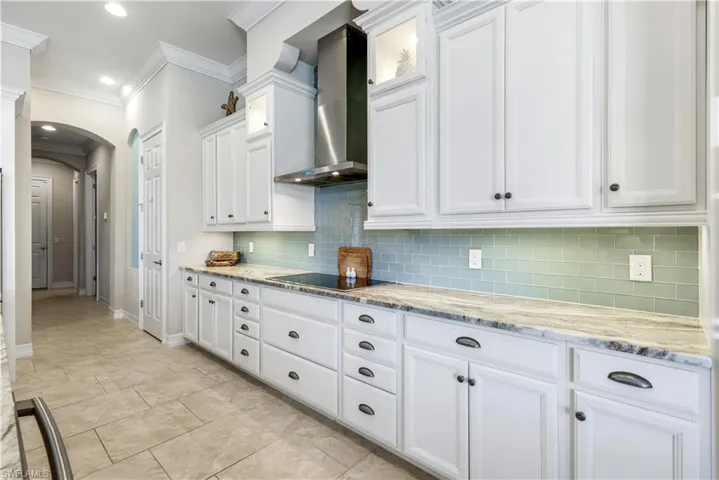 Kitchen featuring white cabinets, arched walkways, light stone counters, wall chimney range hood, and backsplash