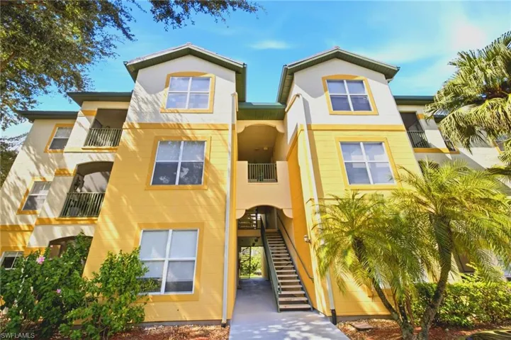 Multi-story building exterior featuring a stucco facade, dual gabled rooflines, recessed balconies with metal railings, and an open-air staircase