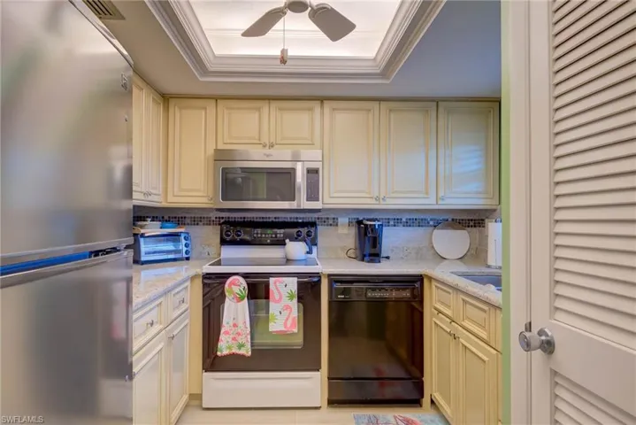 Kitchen with cream cabinetry, appliances with stainless steel finishes, a raised ceiling, light stone counters, and ornamental molding