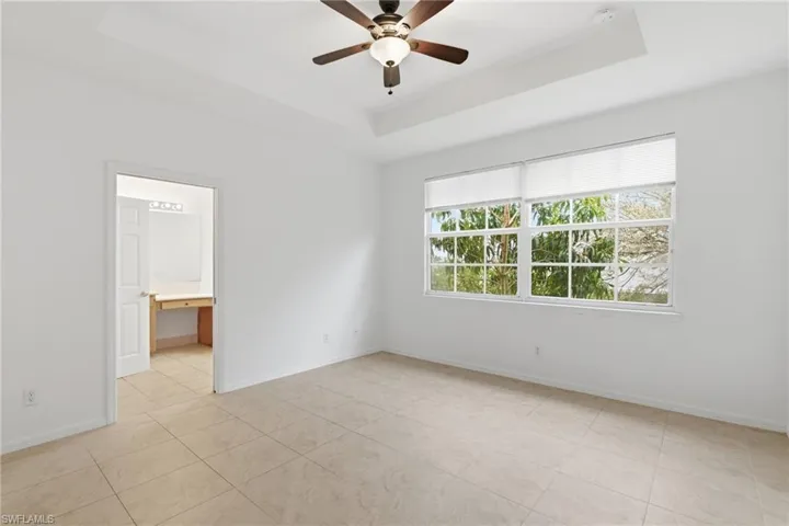 Unfurnished bedroom with a raised ceiling, ceiling fan, built in desk, light tile patterned flooring, and ensuite bathroom