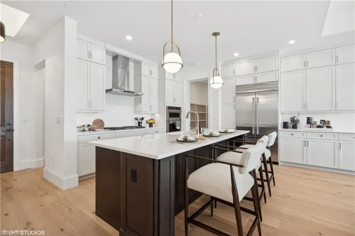 Kitchen featuring pendant lighting, a center island with sink, wall chimney range hood, light hardwood / wood-style flooring, and appliances with stainless steel finishes