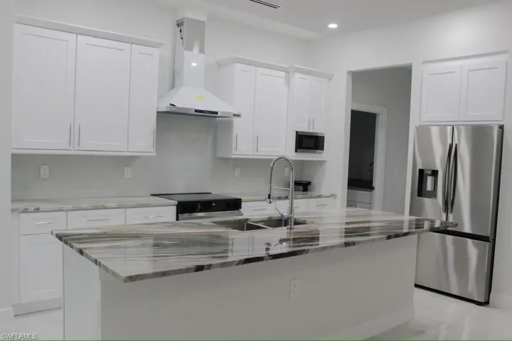 Kitchen with white cabinetry, an island with sink, light stone counters, wall chimney range hood, and appliances with stainless steel finishes