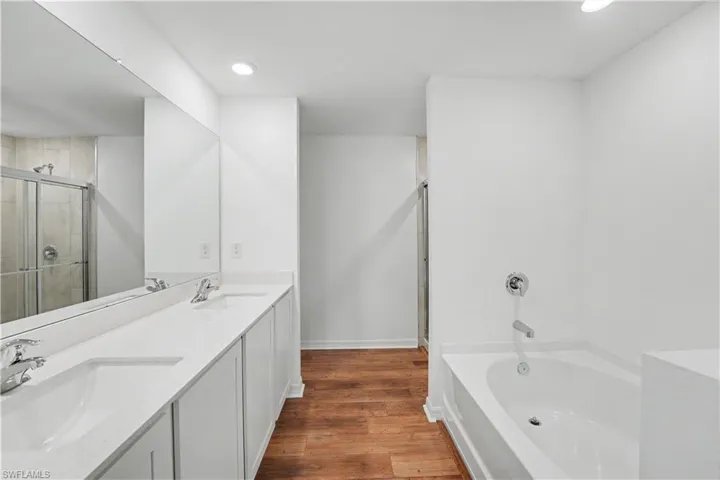 Spacious bathroom featuring a dual vanity with white cabinetry and a continuous countertop, a built-in bathtub with chrome fixtures, a separate glass-enclosed shower, and wood-finish flooring