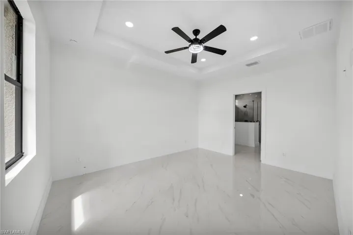 Unfurnished bedroom featuring a tray ceiling, ceiling fan, recessed lighting, light marble finish floors, and ensuite bath