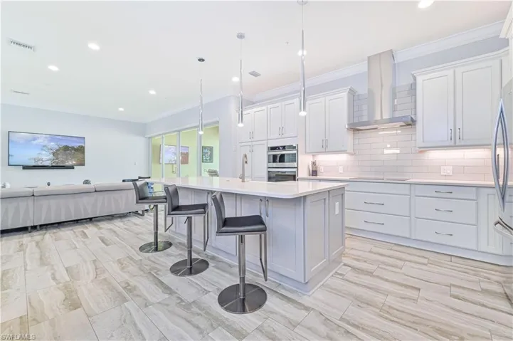 Kitchen with a breakfast bar area, white cabinetry, hanging light fixtures, wall chimney exhaust hood, and a center island with sink