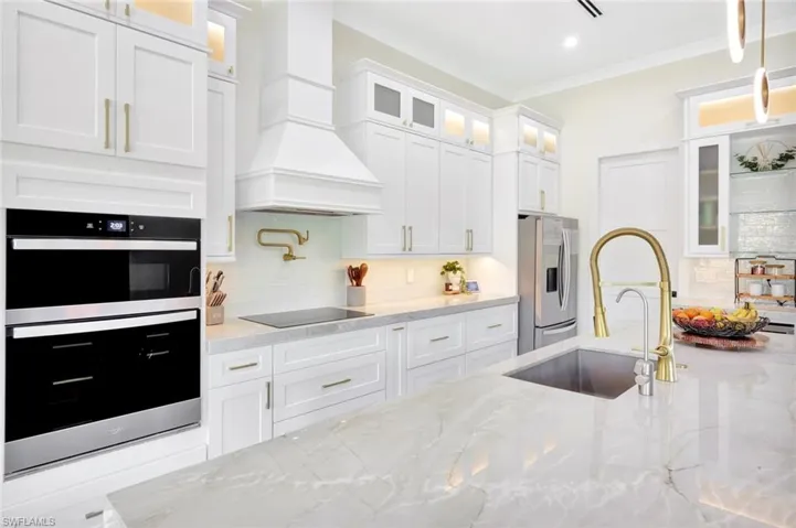Kitchen with stainless steel appliances, glass insert cabinets, backsplash, light stone countertops, and white cabinetry