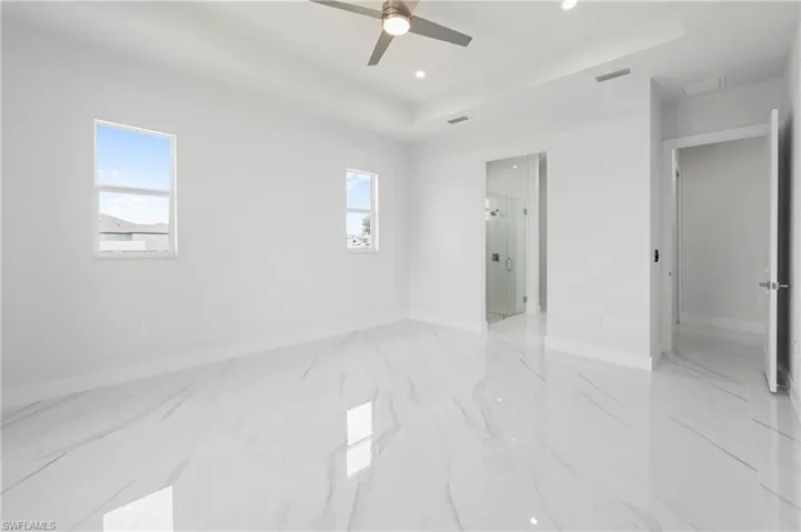 Spacious room featuring large format tile flooring, a tray ceiling with recessed lighting and a ceiling fan, dual windows, and an en-suite bathroom with a glass-enclosed shower