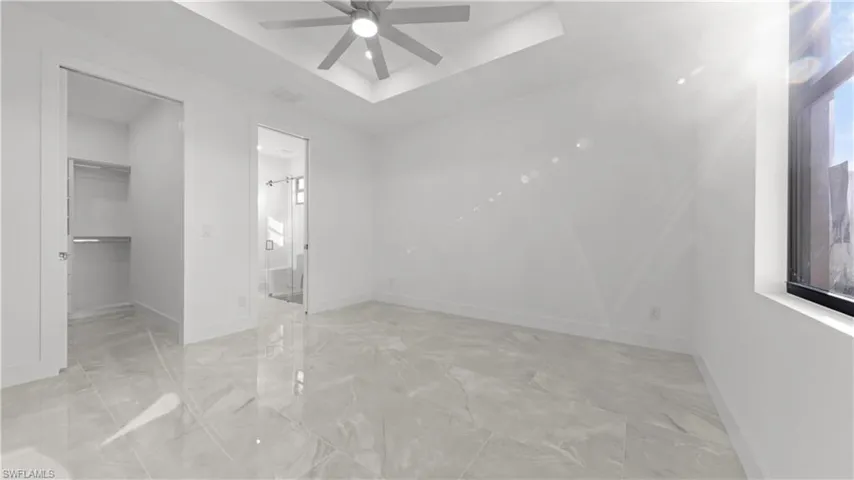 Unfurnished bedroom with a tray ceiling, a spacious closet, ceiling fan, light marble finish floors, and ensuite bathroom