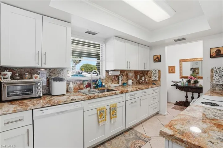 Kitchen featuring a tray ceiling, ornamental molding, white dishwasher, light stone counters, and white cabinetry