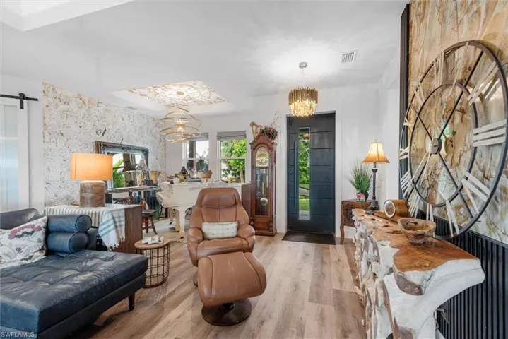 Spacious living area featuring wood-finish flooring, a contemporary chandelier, a unique textured accent wall, and an entryway with a dark paneled door and sidelight window