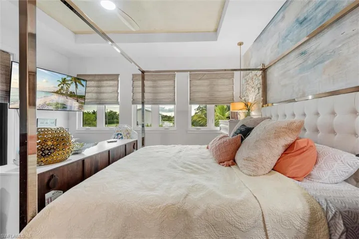 Recessed ceiling with integrated ceiling fan, multiple windows with neutral-toned Roman shades, built-in storage cabinetry, tufted upholstered headboard, and a unique textured wall treatment