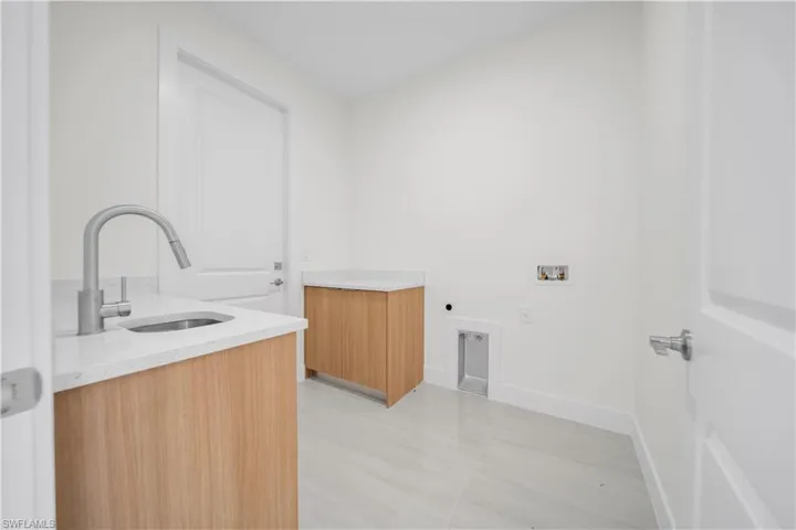 Laundry area featuring light wood-finish cabinetry, light-toned countertops, a stainless steel sink with a pull-down faucet, and light-toned tile flooring