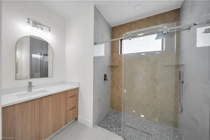 Contemporary bathroom featuring a wood-finish vanity with a white countertop and rectangular sink, a frameless shower with mosaic tile flooring, a horizontal accent window, and a patterned tile shower wall