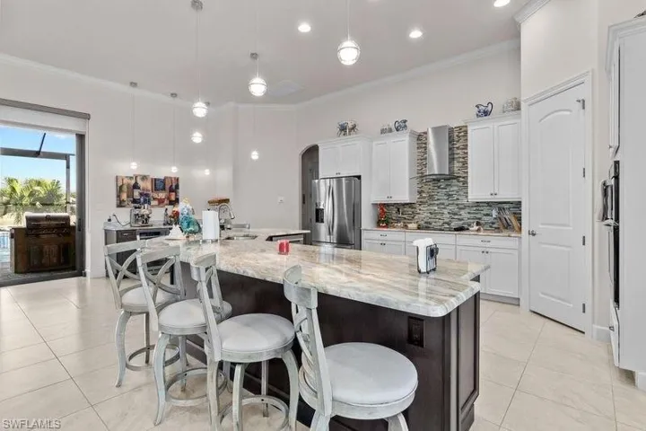 Kitchen featuring a spacious island, wall chimney range hood, decorative light fixtures, stainless steel fridge with ice dispenser, and white cabinetry