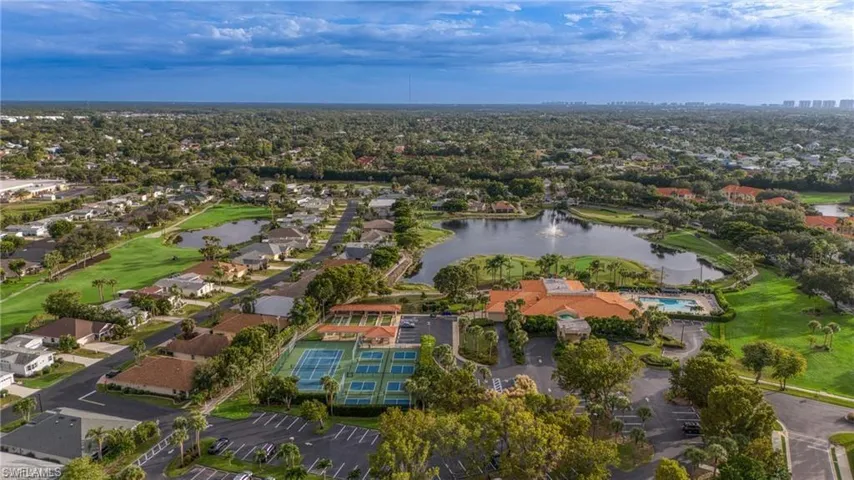 Aerial perspective showcasing a vibrant community with a central lake featuring a fountain, multiple tennis courts, and a club building with an outdoor swimming pool