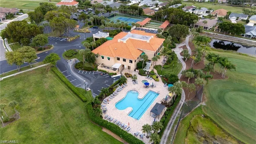 Community clubhouse featuring a terracotta tile roof, a large freeform swimming pool with integrated lap lane, an expansive parking area, and tennis courts