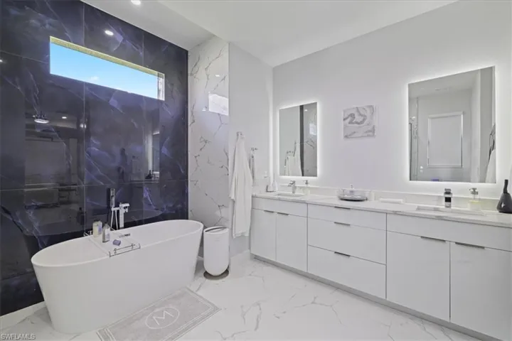 Full bath featuring light marble finish flooring, a soaking tub, double vanity, and stone wall