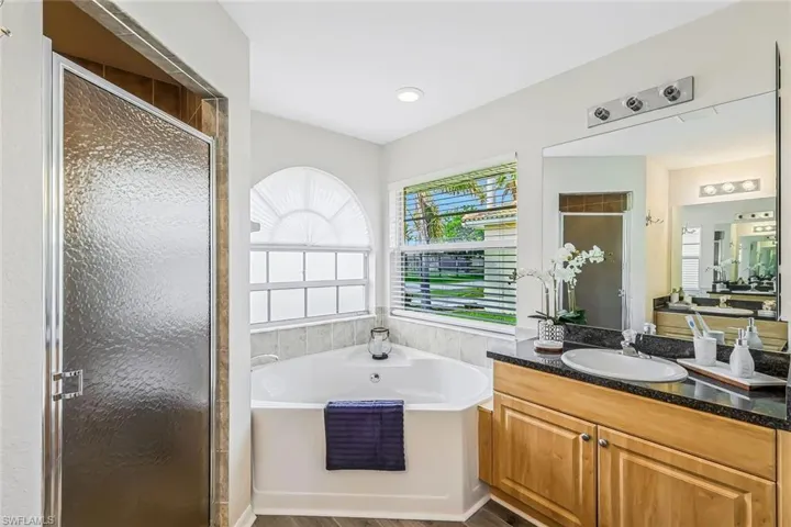 Spacious bathroom featuring a corner soaking tub with arched window, a stand-up shower with textured glass door, a vanity with wood-finish cabinetry, a dark countertop, and an undermount sink