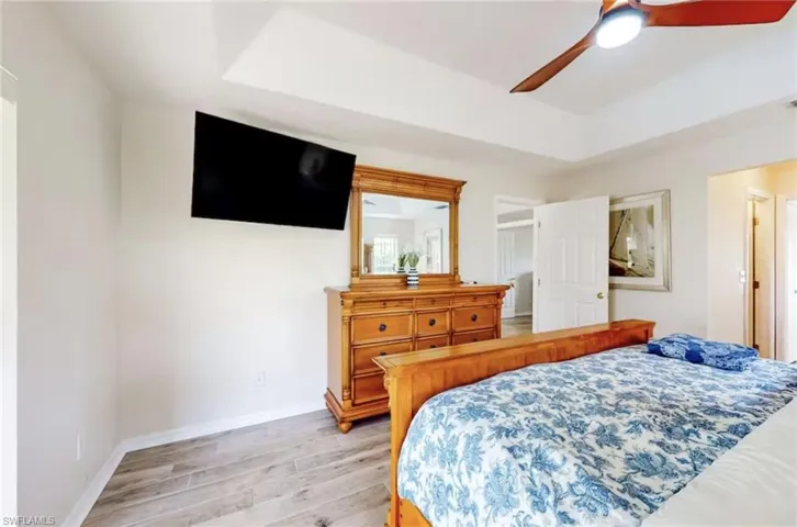 Bedroom featuring tray ceiling, wood-finish flooring, ceiling fan with integrated lighting, wall-mounted television, and a white paneled door