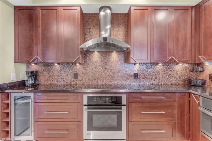 Kitchen with stainless steel oven, wall chimney exhaust hood, beverage cooler, and dark stone counters